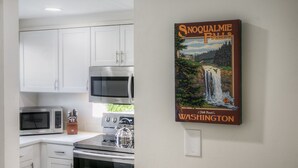 A kitchen with white cabinets, a microwave, a kettle, and a poster of Snoqualmie Falls. - Moon River Suites 1 - on River, Private Hot Tub, No Flood Impact (North Bend)