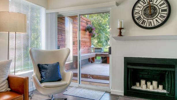 A modern living room with a fireplace, a large wall clock, and a sliding glass door leading to an outdoor deck. - Moon River Suites 1 - on River, Private Hot Tub, No Flood Impact (North Bend)