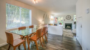 A modern living room with a glass dining table, wooden chairs, a fireplace, and a large window with blinds. - Moon River Suites 1 - on River, Private Hot Tub, No Flood Impact (North Bend)