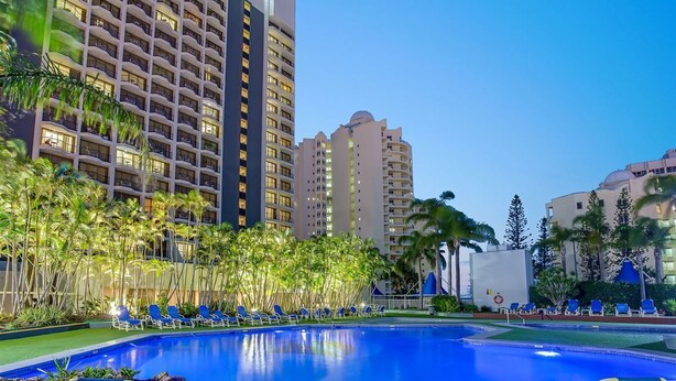 Zona de piscina de hotel con múltiples sillas de descanso, palmeras y edificios altos al fondo.
