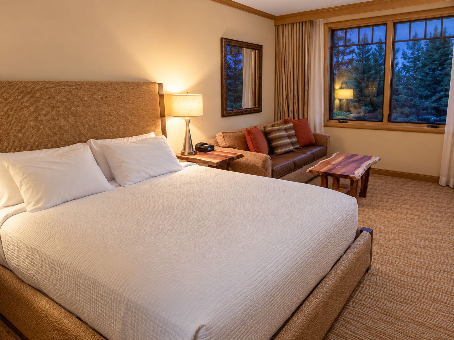 A hotel room with a large bed, a bedside table with a lamp, a sofa, a small table, and a window with a view of trees.