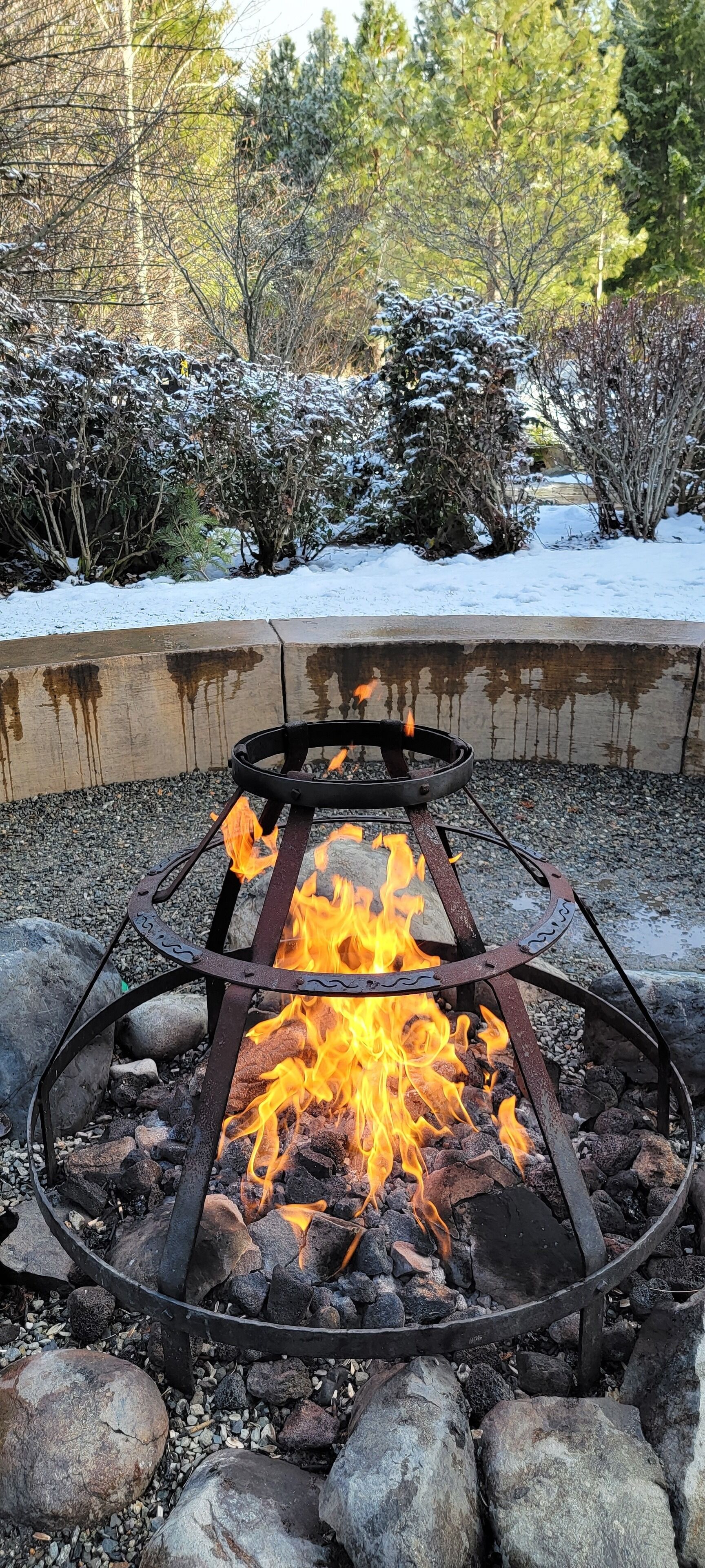A metal fire pit with flames, surrounded by rocks and snow-covered plants.