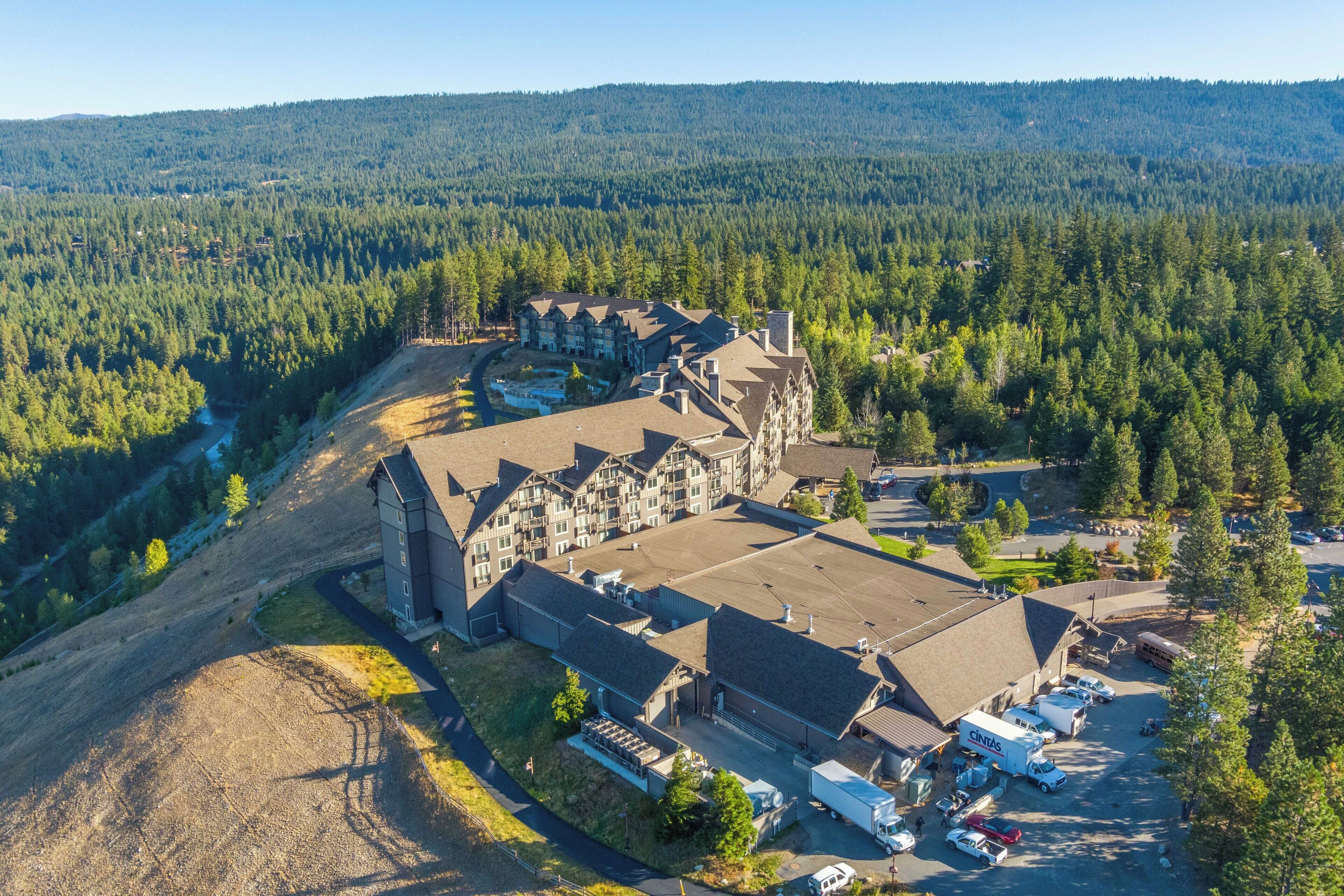 Une vue aérienne d’un vaste complexe de bâtiments entouré d’une forêt dense, avec plusieurs aires de stationnement et une route bien visible menant à l’entrée.