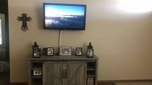 A living room with a mounted TV, a wooden cabinet with framed photos, a cross on the wall, and a sign that reads "FAITH FAMILY FRIENDS". - Cute, cozy, close duplex. (Stillwater)