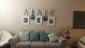 A living room with a beige sofa, teal and green cushions, a side table with a lamp, and a wall-mounted picture frame holder with several framed photos. - Cute, cozy, close duplex. (Stillwater)