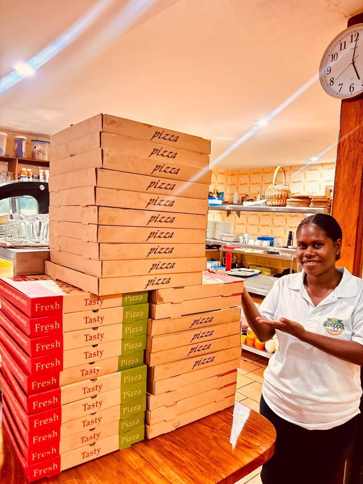 A person standing next to a stack of pizza boxes labeled 'Fresh' and 'Tasty' in a kitchen setting with a clock on the wall.