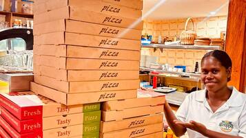 A person standing next to a stack of pizza boxes labeled 'Fresh' and 'Tasty' in a kitchen setting with a clock on the wall.
