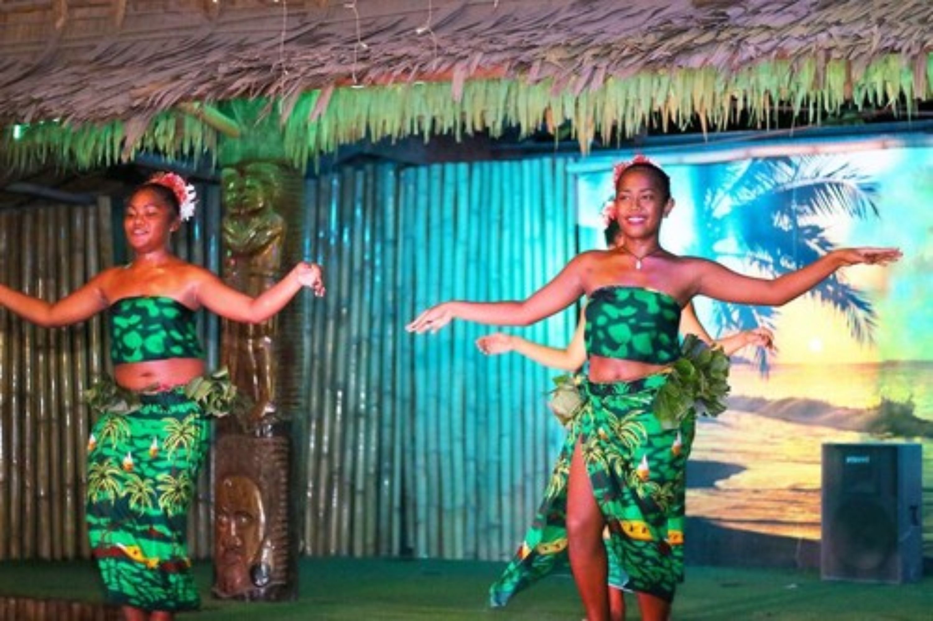 A stage with two performers in green tropical costumes, a backdrop with palm trees, and a thatched roof structure.