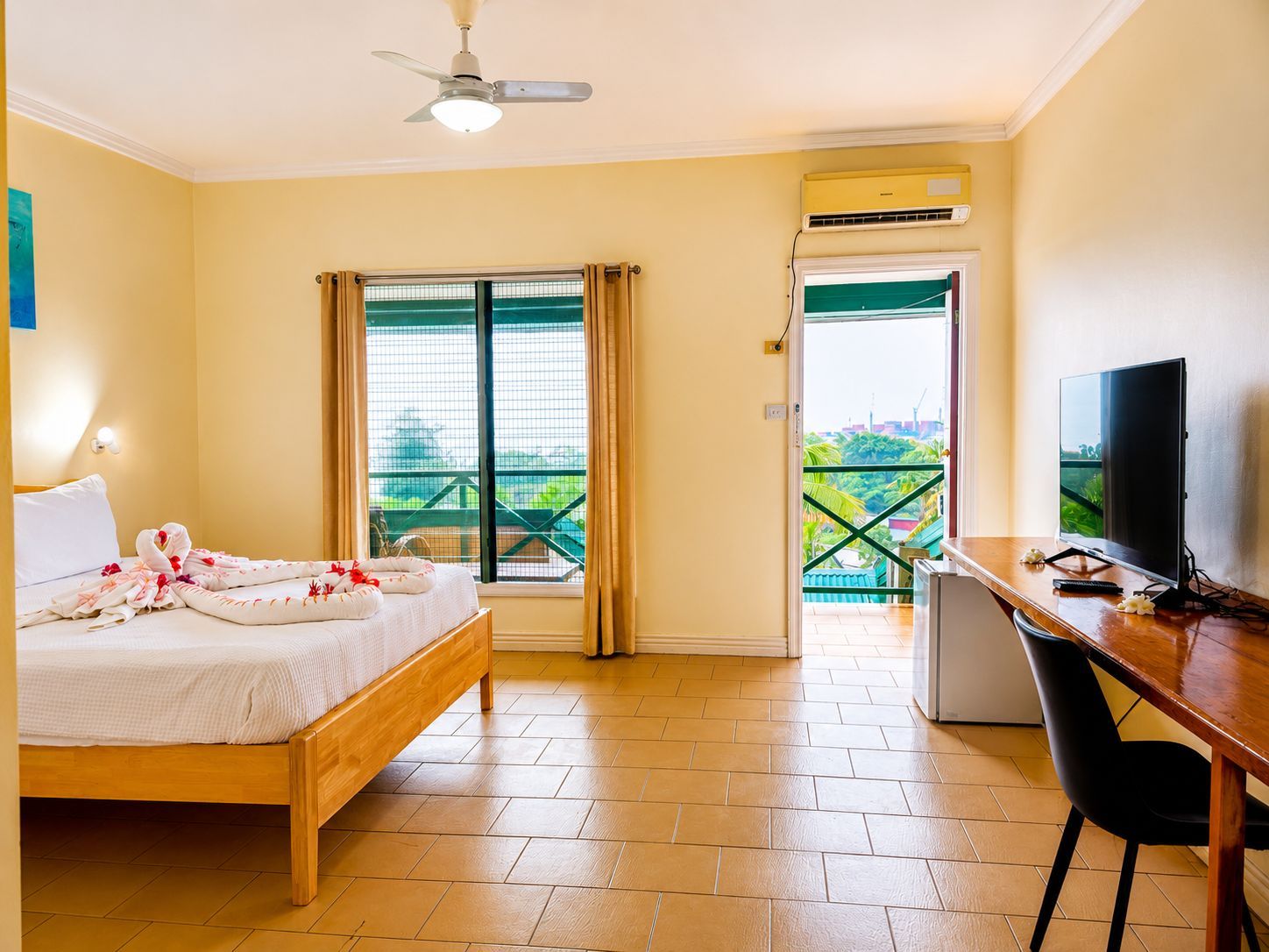 A hotel room with a bed, a desk with a TV, an air conditioning unit, and a balcony with a view.