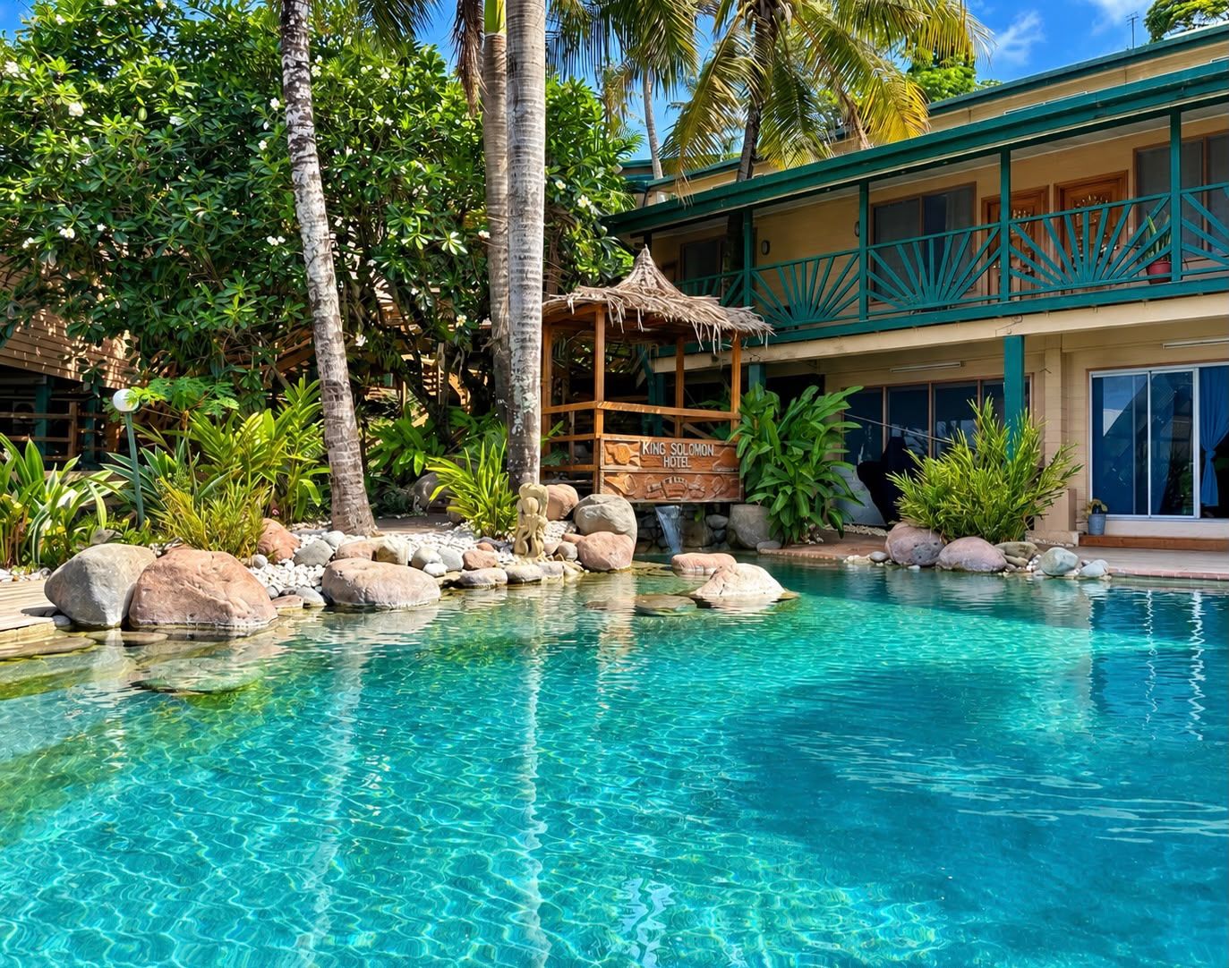 A clear pool with rocks and a wooden structure, surrounded by lush greenery and a building with a balcony.