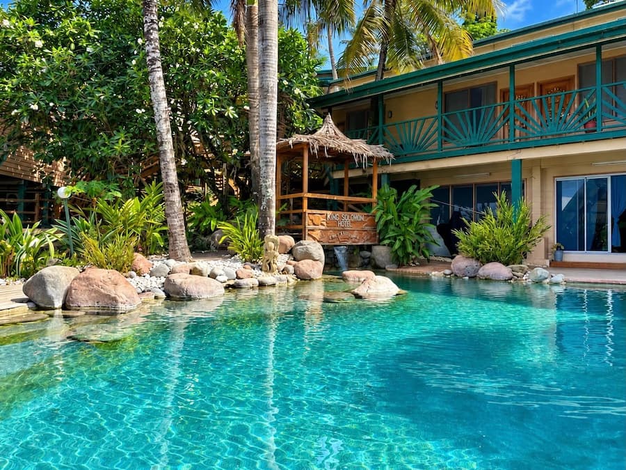 A clear pool with rocks and a wooden structure, surrounded by lush greenery and a building with a balcony.