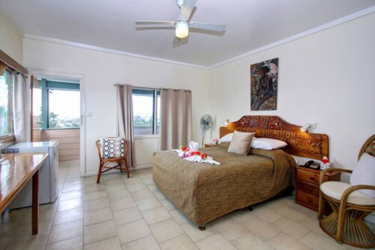 A bedroom with a bed, a desk, a chair, a ceiling fan, and a window.