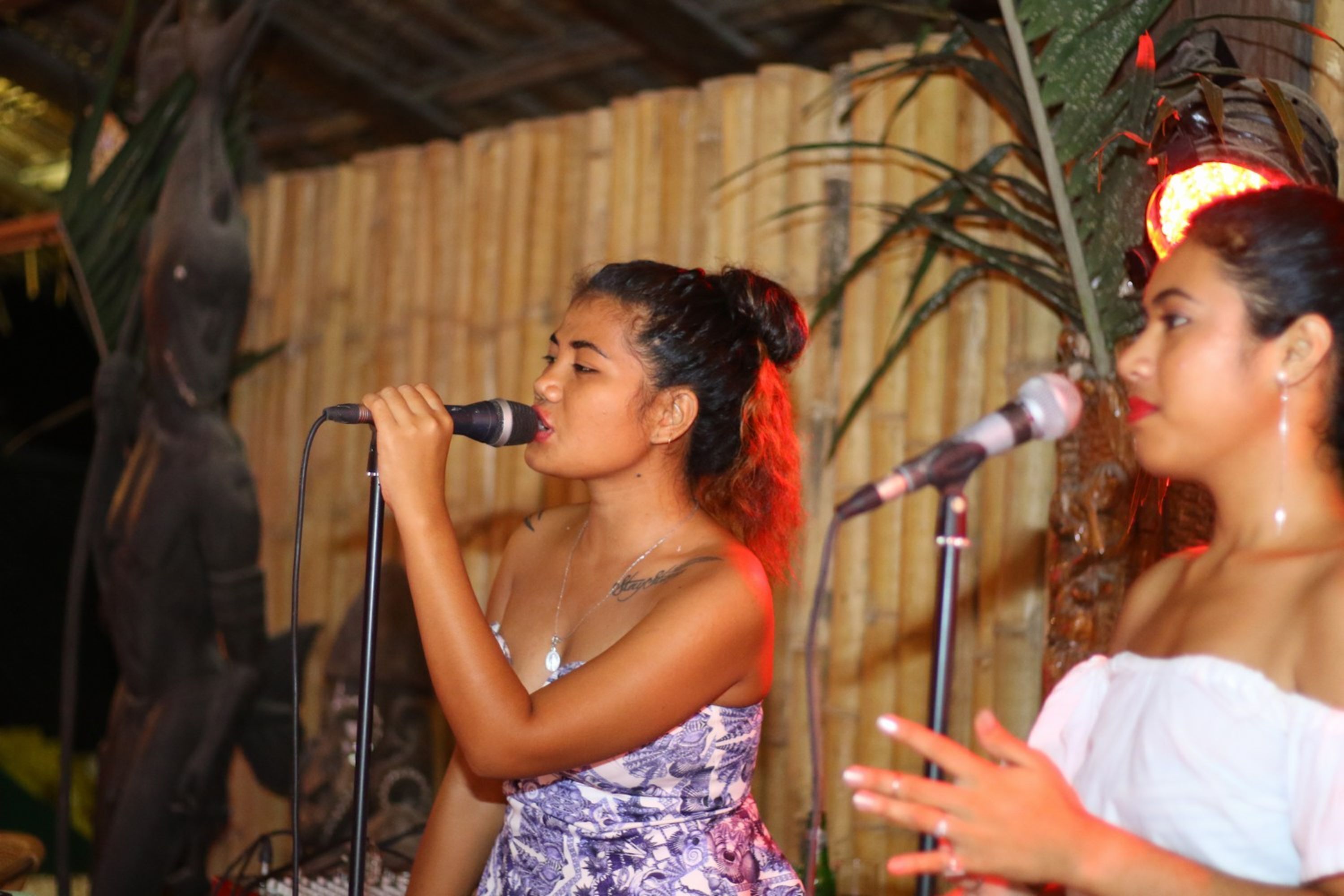 Two individuals performing on stage with microphones, surrounded by bamboo structures and tropical plants.