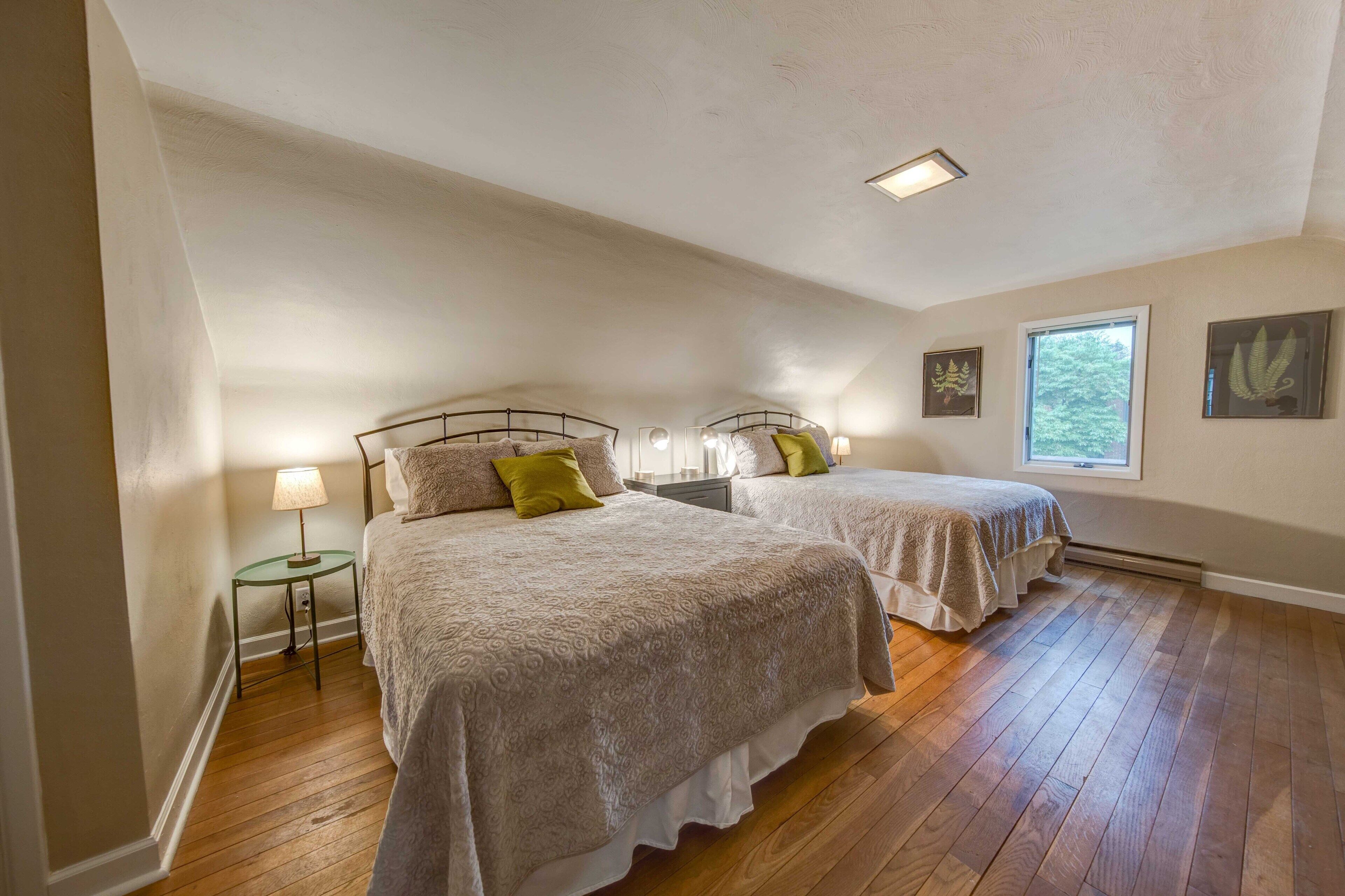 A bedroom with two beds, a small table with a lamp, and a window with a view of trees.