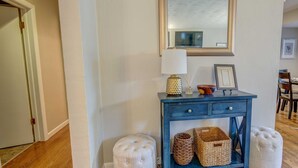 Een gang met een blauwe console tafel, een spiegel en twee witte poefs.