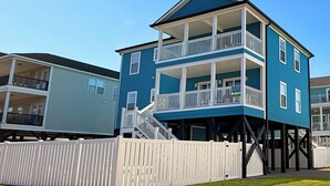 Une maison de deux étages située en bord de mer, dotée d’un balcon à l’étage supérieur, d’une clôture blanche en bois et surplombant un ciel d’un bleu éclatant.