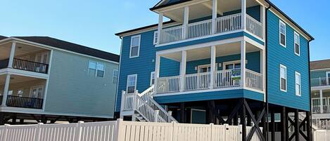 A two-story beachfront house with a balcony on the upper floor, a white picket fence, and a clear blue sky.