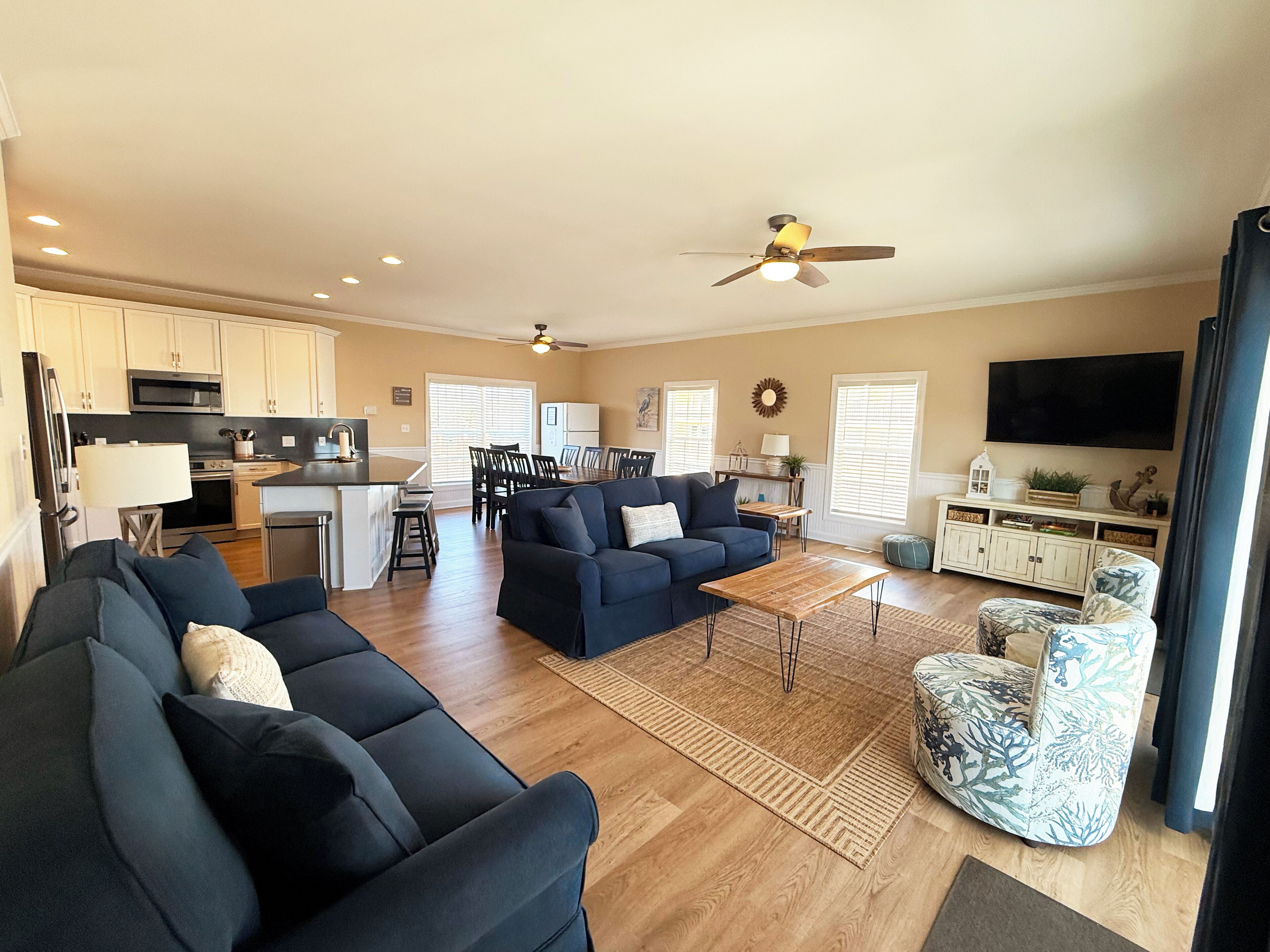 A spacious living area with a sofa set, a coffee table, and a patterned armchair. A kitchen with white cabinets and a dining area with a table and chairs are visible in the background.