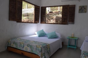 Ein Schlafzimmer mit einem Bett, zwei Fenstern mit Holzläden, einem kleinen Tisch mit einem dekorativen Gegenstand und einem an der Wand hängenden Bild.