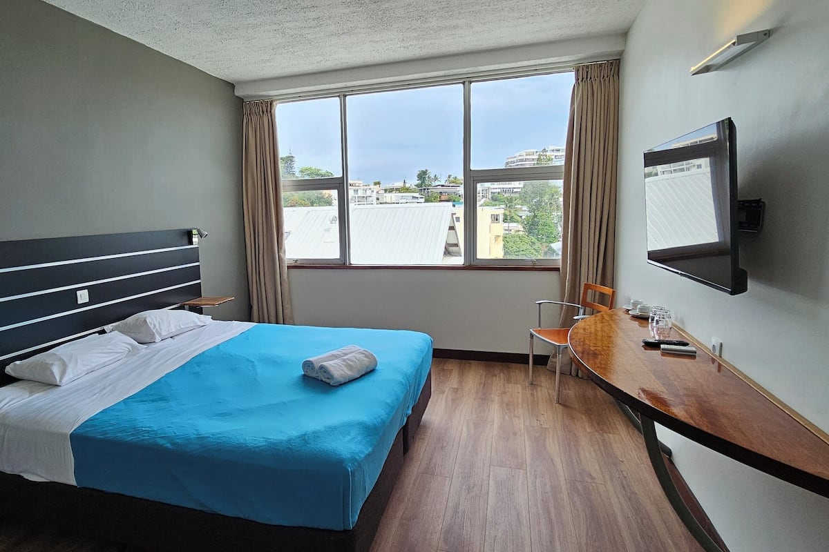 Quarto de hotel com cama, escrivaninha, cadeira, televisão e janela com vista para edifícios.