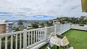 Ein Balkon mit weißer Reling, eine kleine Rasenfläche und Blick auf einen Hafen sowie Gebäude in der Ferne.