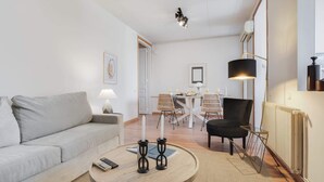 A modern living room with a grey sofa, a round wooden coffee table, and a black armchair. There is a dining table with chairs in the background, a floor lamp, and a ceiling light fixture. - Sweett | Rocafort 404 (Barcelona)