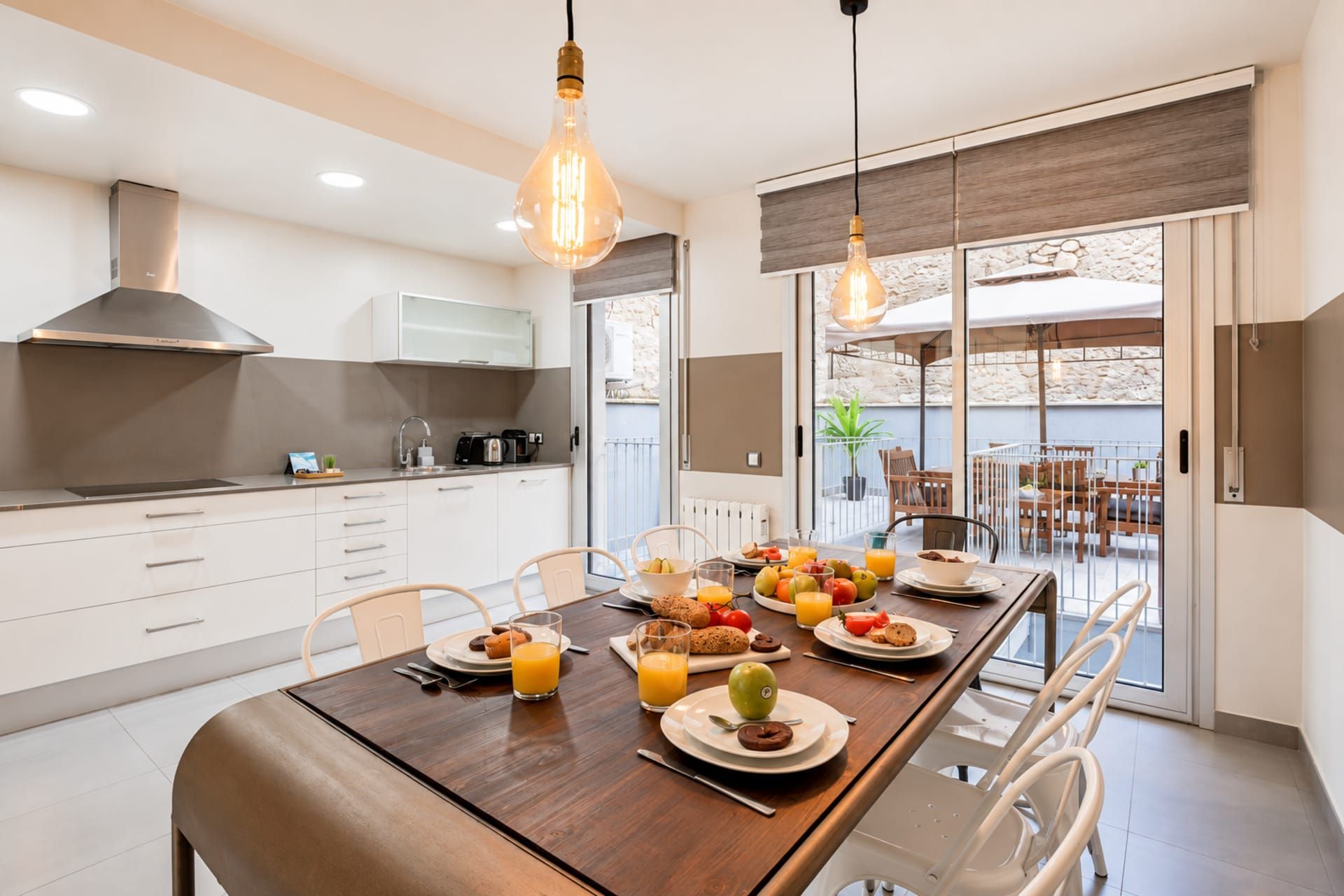 Une cuisine moderne avec un coin repas, comprenant une table en bois dressée pour le petit-déjeuner, avec divers fruits et viennoiseries.