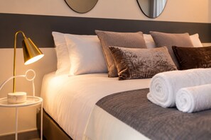 Een opgemaakt bed met witte en bruine kussens, een gouden lamp op een witte nachtkastje, en een opgerolde witte handdoek op het bed.