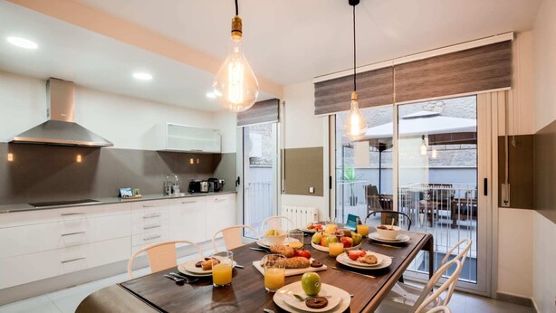 Une cuisine moderne avec un coin repas, où une table en bois est dressée pour le petit-déjeuner, avec des fruits, des viennoiseries et du jus d'orange.