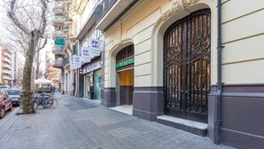 Eine Straße mit einer Reihe von Geschäften, darunter ein 'Mercado' und ein Schild für 'Mediterranean Marketing', und ein verschlossener Eingang mit schmiedeeisernem Zierwerk.