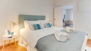 Ein ordentlich bezogenes Bett mit weißen und gemusterten Kissen, einer blauen Tagesdecke und aufgerollten Handtüchern auf einer weißen Bettdecke. Ein kleiner weißer Nachttisch mit Lampe und Flasche ist zu sehen. Der Raum hat weiße Wände und Holzboden.