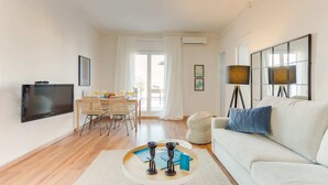 Ein modernes Wohnzimmer mit Flachbildfernseher, einem Essbereich mit Tisch und Stühlen, einem Sofa mit blauem Kissen und einer Lampe.