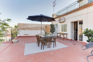 Une terrasse avec une table, des chaises et un parasol. On y trouve une clôture en bois, des plantes en pot et un bâtiment doté d’un balcon.