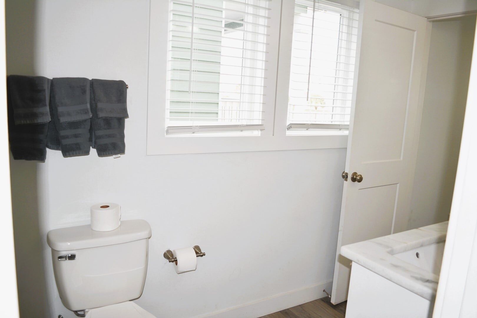 Een badkamer met een toilet, twee rollen toiletpapier, een handdoekhouder met handdoeken en een raam met zonwering.