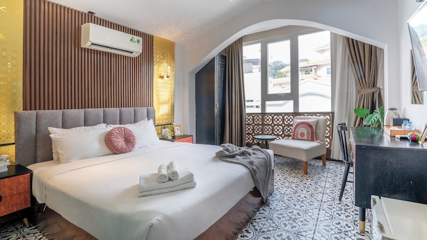A hotel room with a large bed, an air conditioner, a desk with a chair, a small table, and a view of the outside.