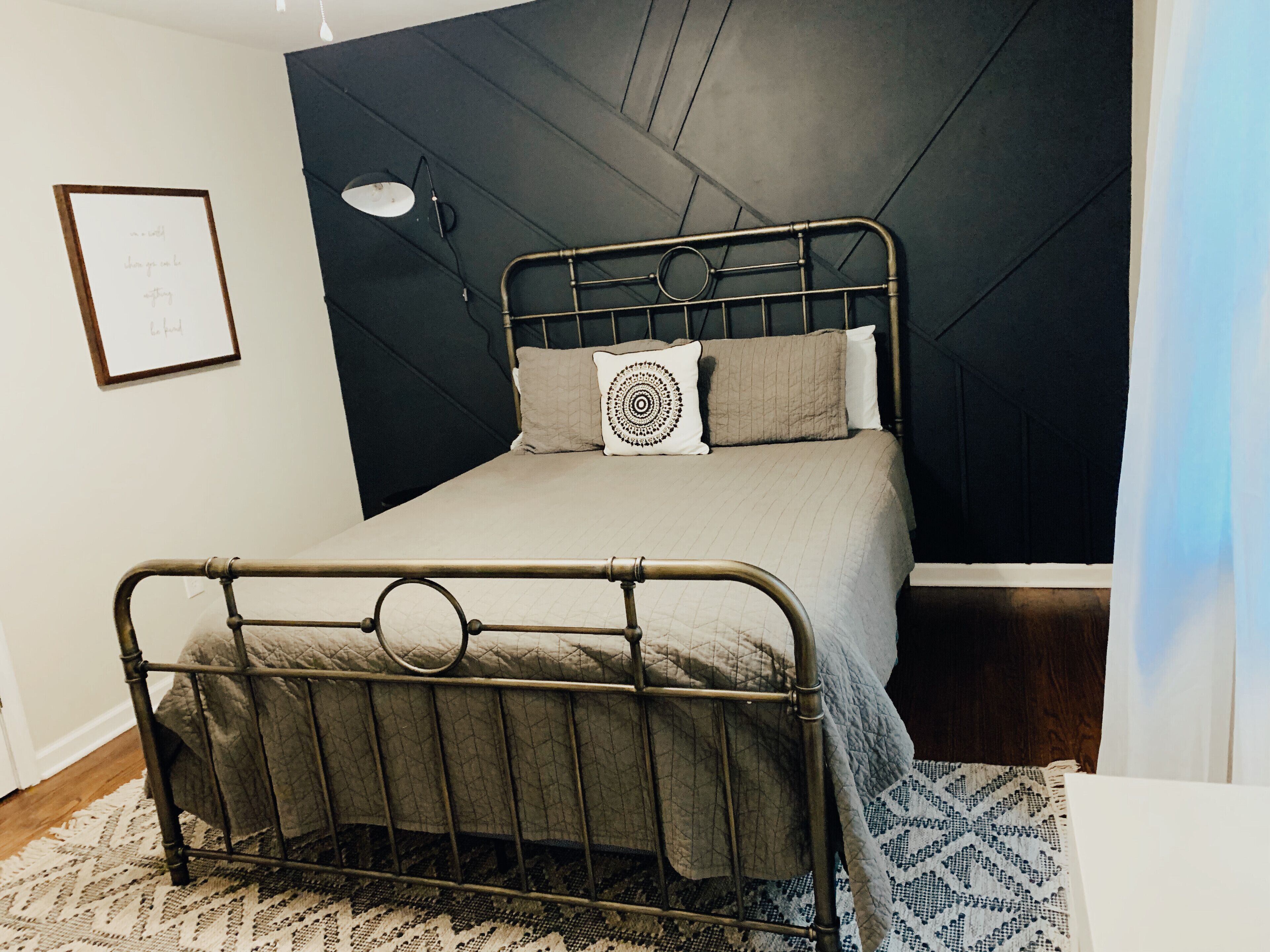 A bed with a metal frame, a quilted mattress, and two pillows. There is a wall-mounted lamp and a framed picture on the wall.