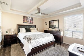 Une chambre à coucher avec un grand lit, une commode, une table de chevet, un ventilateur de plafond et une fenêtre avec des stores.