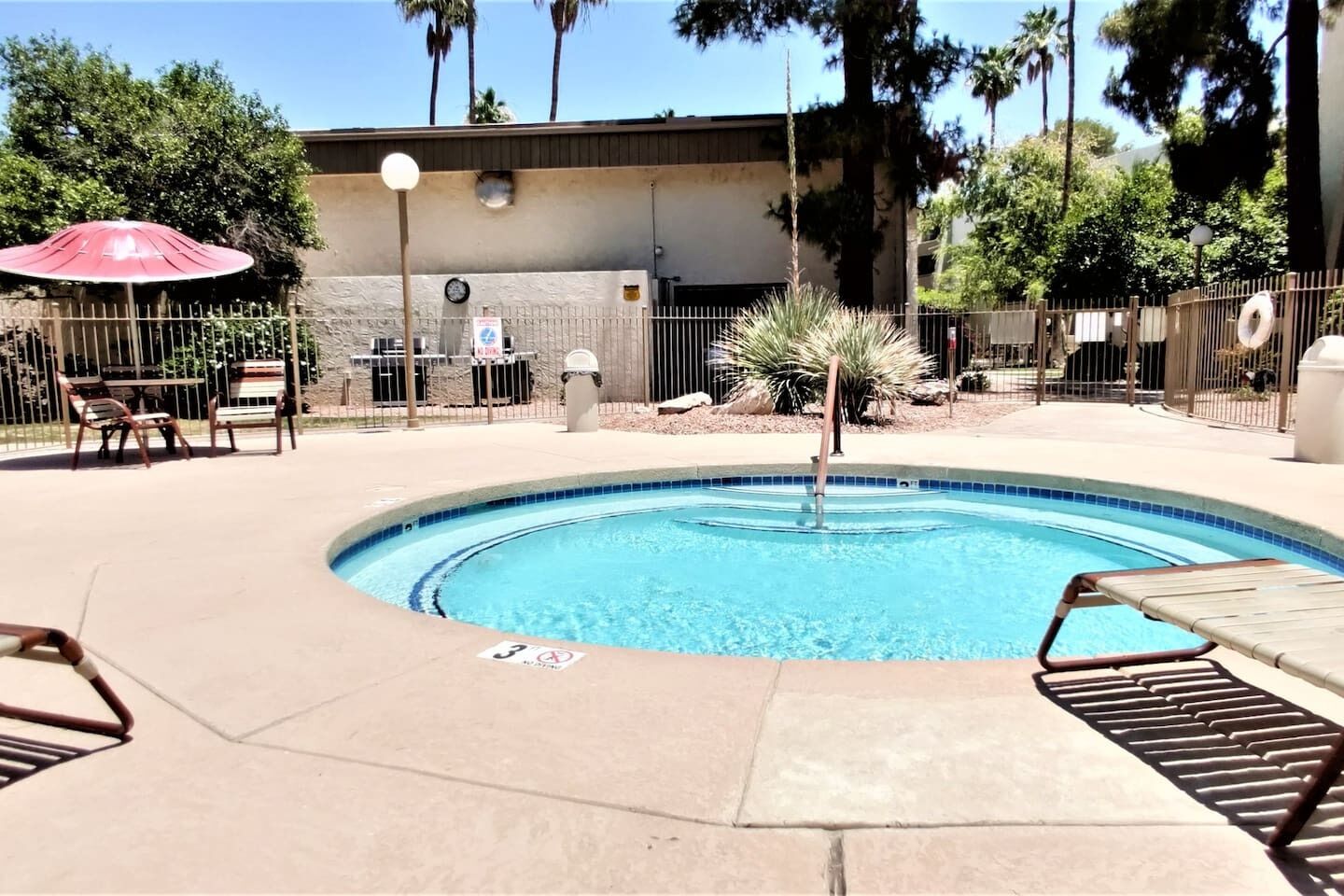 A swimming pool with a red umbrella, lounge chairs, and a fenced area with trees and a building in the background.