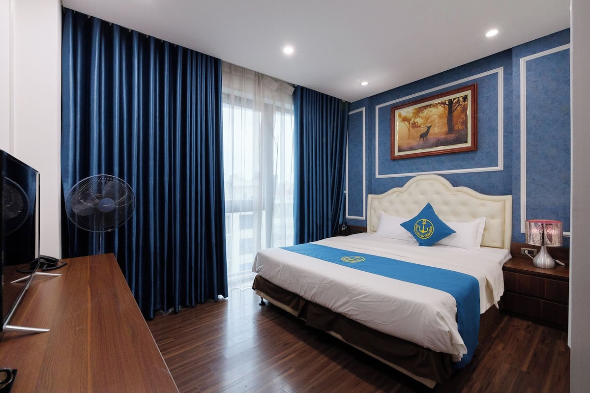 A hotel room with a bed, a desk, a fan, a painting, and a lamp.