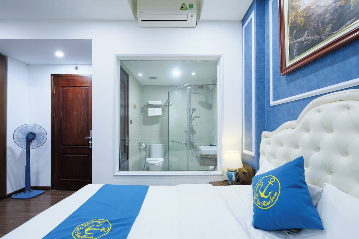 A hotel room with a bed, a fan, a glass-enclosed shower, and a painting on the wall.