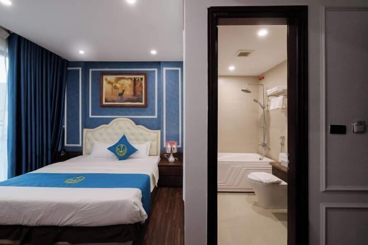 A hotel room with a bed, a nightstand, a painting on the wall, and a bathroom visible through an open door.