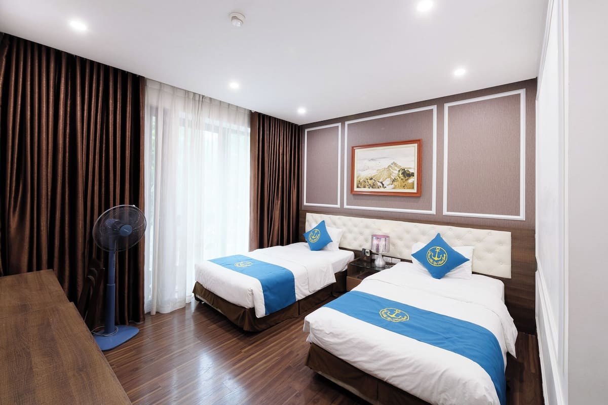 A hotel room with two beds, a wooden floor, a fan, a desk, and a painting on the wall.