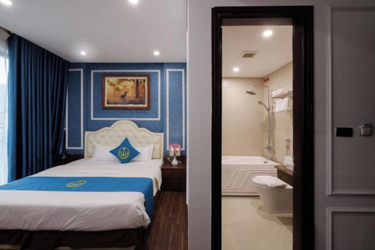 A hotel room with a bed, a nightstand, a painting on the wall, and a bathroom visible through an open door.