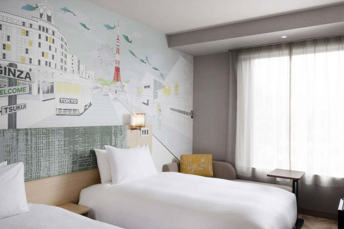 Une chambre d’hôtel avec deux lits, une fresque représentant un paysage urbain sur le mur, une table de chevet, une lampe et une vue sur l’extérieur par une fenêtre.