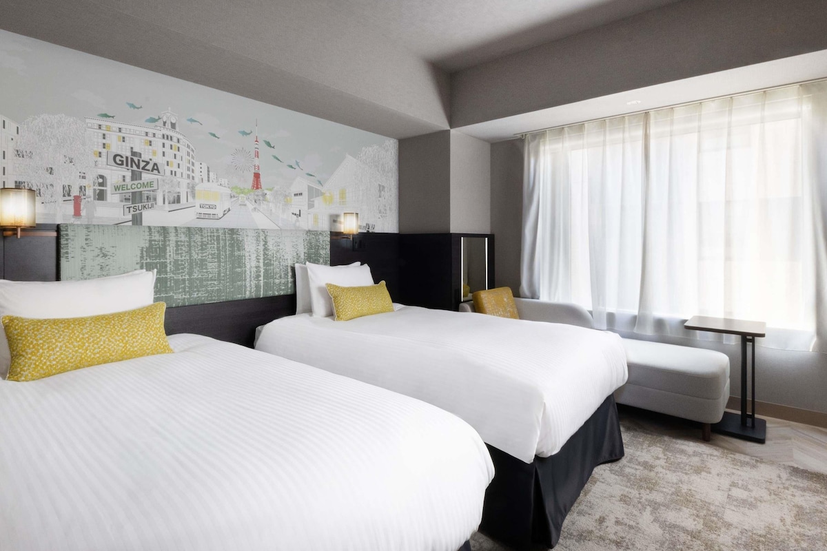 Een hotelkamer met twee bedden, een groot stadsgezicht muurschildering, een bureau en een raam met dunne gordijnen.