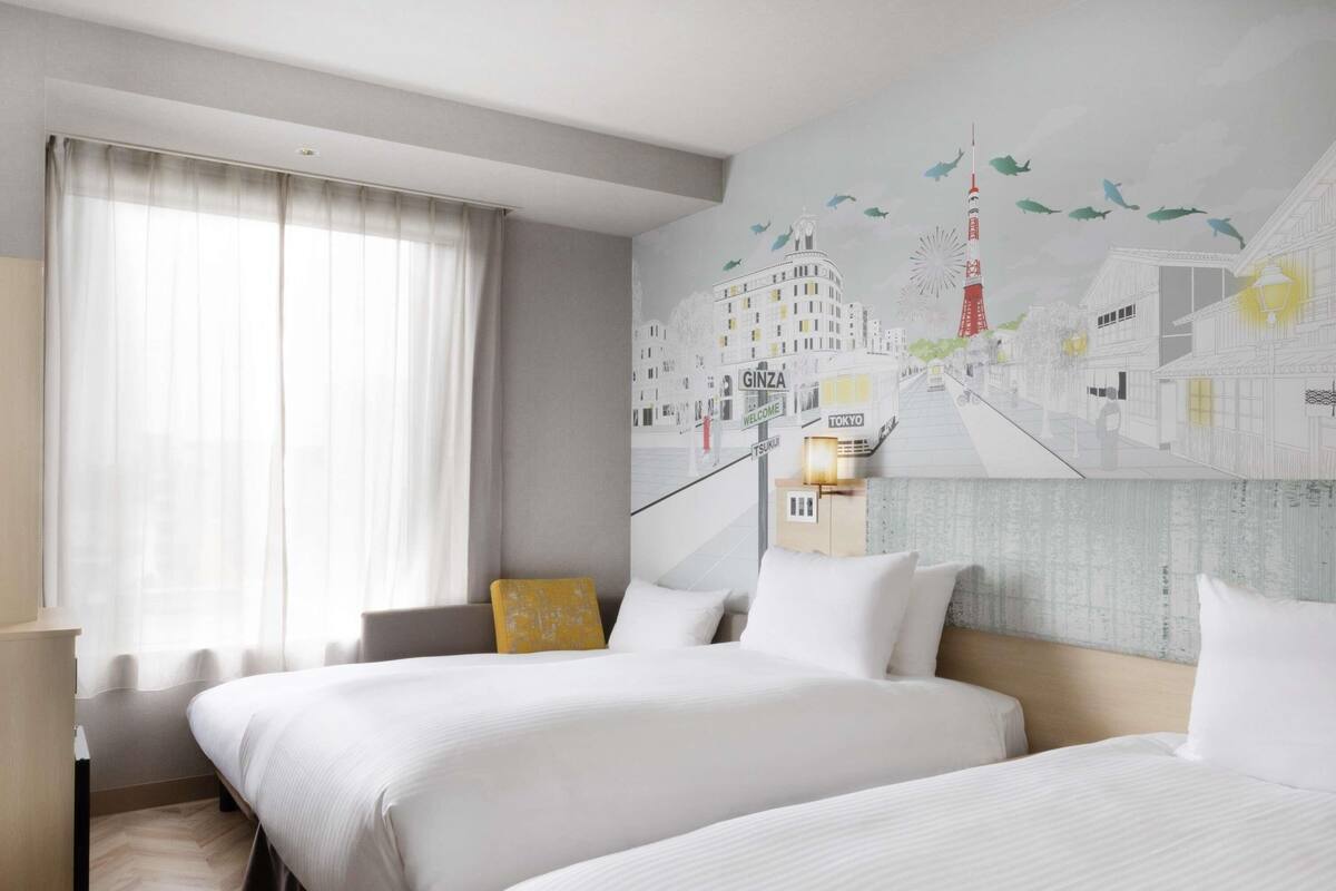 Une chambre d’hôtel avec deux lits, une grande fenêtre avec des rideaux, une fresque représentant un paysage urbain sur le mur et une table de chevet avec une lampe.