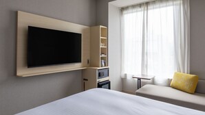 Habitación de hotel con un televisor de pantalla plana grande montado en la pared, una cama con colchón blanco, una mesita de noche pequeña, un sofá con un cojín amarillo y una ventana con cortinas traslúcidas.
