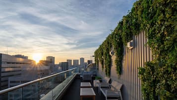 A rooftop terrace with a glass balustrade, outdoor seating, and a city skyline view during sunset.