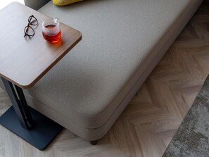Un sofá con tela texturizada, una mesita de madera con un par de gafas y un vaso de líquido rojo, y un suelo de madera con patrón de espiga.