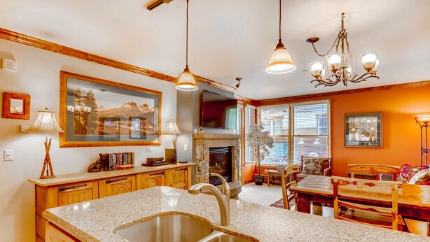 A kitchen with granite countertops, a stainless steel sink, wooden cabinets, a stone fireplace, and a dining area with wooden furniture. - Updated 1br Premier  At Black Bear Lodge- Kids Ski Free 1 Bedroom Condo by RedAwning (Keystone)